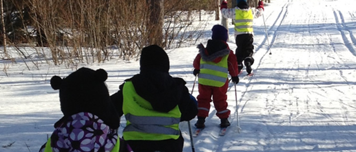 Fyra förskolebarn åker skidor i en rad. Gula reflexvästar på.