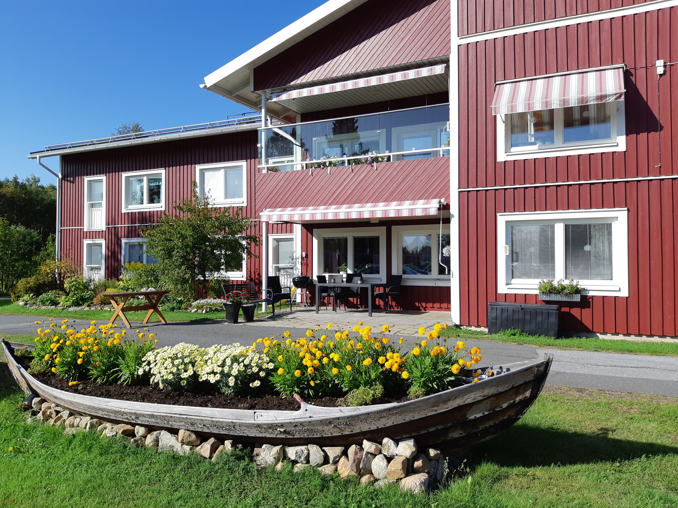 Tallgårdens äldreboende. Båt med blommor, bord och stolar, blommor och buskar utanför huset.