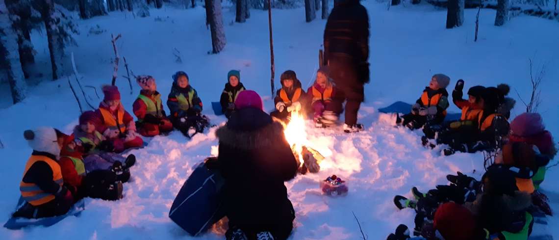 Sjutton förskolebarn sitter runt en eld. Liggunderlag på snön. Två förskollärare berättar något.