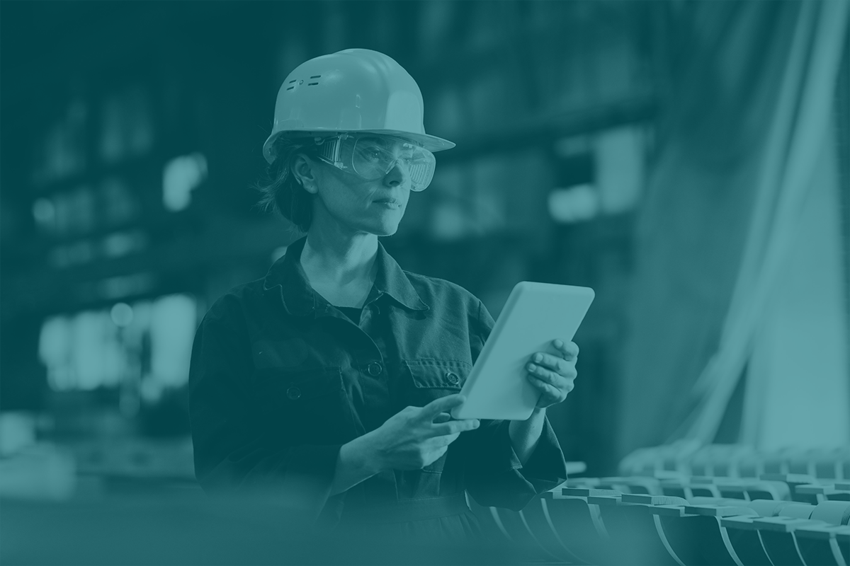 Kvinna med säkerhetshjälm i en industrilokal. Håller i en ipad. Hela bilden täcks av ett grönt filter.